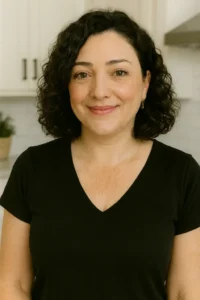 A smiling woman with curly dark hair, wearing a black top, standing in a bright, modern kitchen